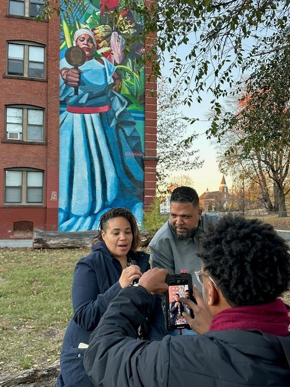A filmmaker recording a community interview near a mural for Beyond the Storm